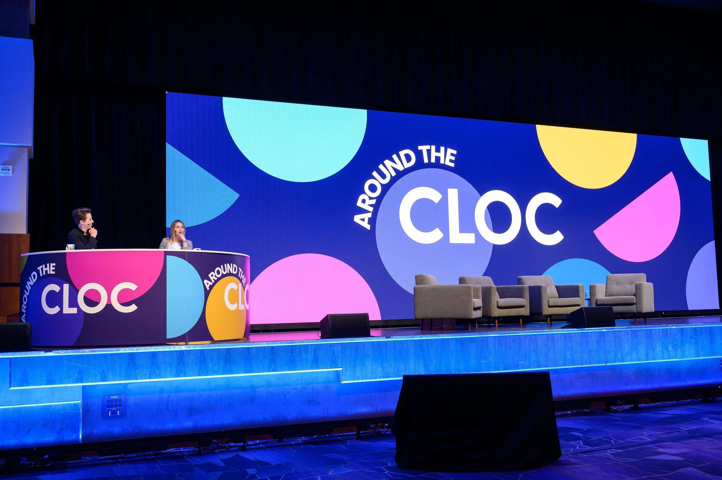 Two presenters at a colorful stage for "Around the CLOC" event with empty gray chairs and a vibrant backdrop.