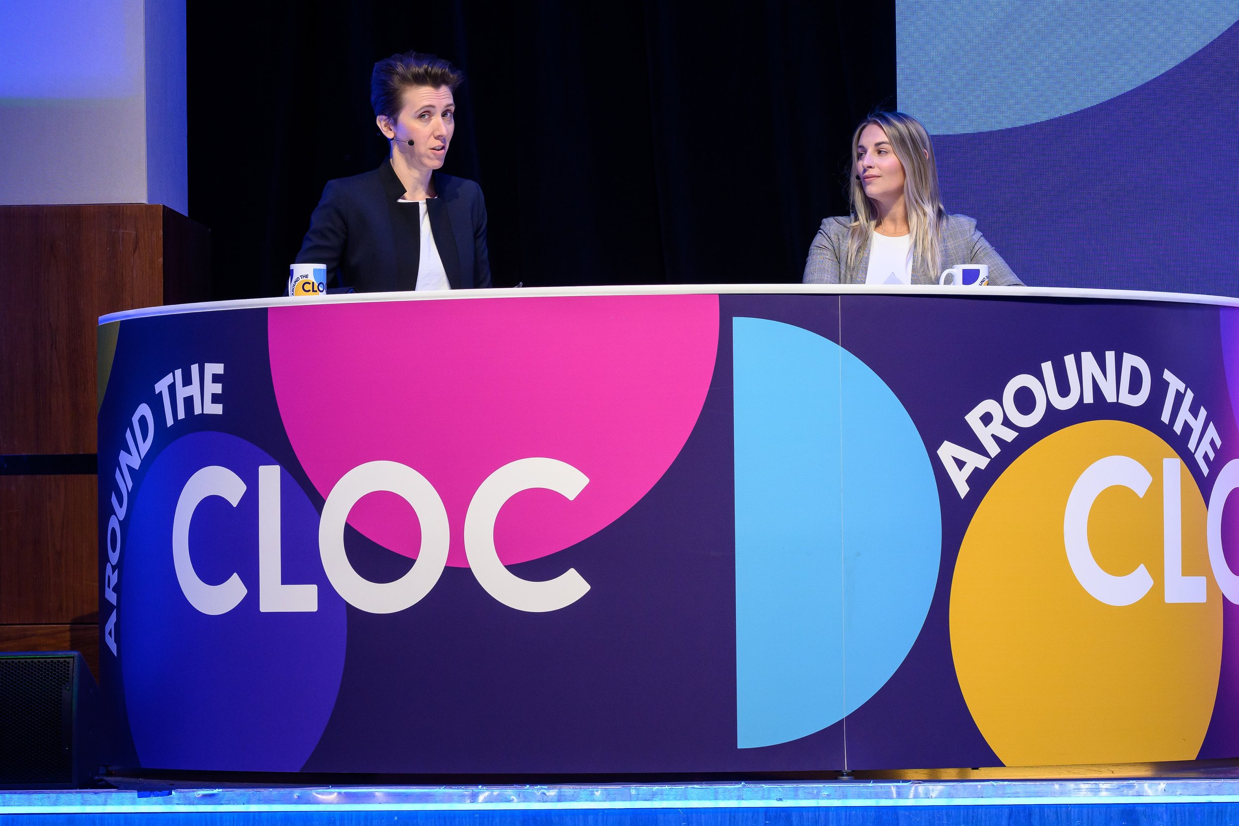 Two people sitting at a desk with colorful "Around the CLOC" branding, engaging in a discussion or presentation. The setting appears to be a conference or event.