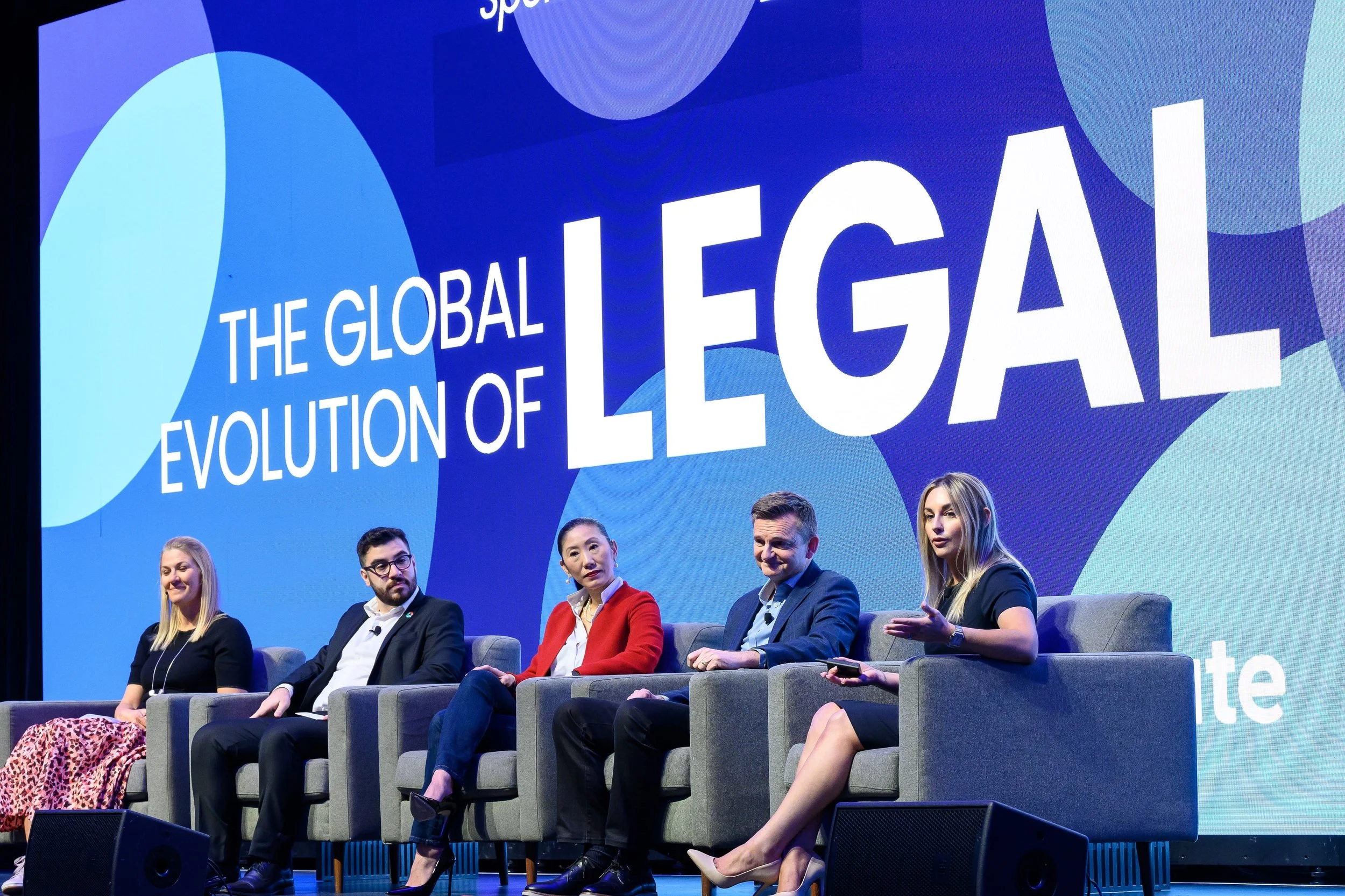 Panel discussion at a legal conference with five speakers seated on stage in front of a blue screen displaying 'The Global Evolution of Legal.'