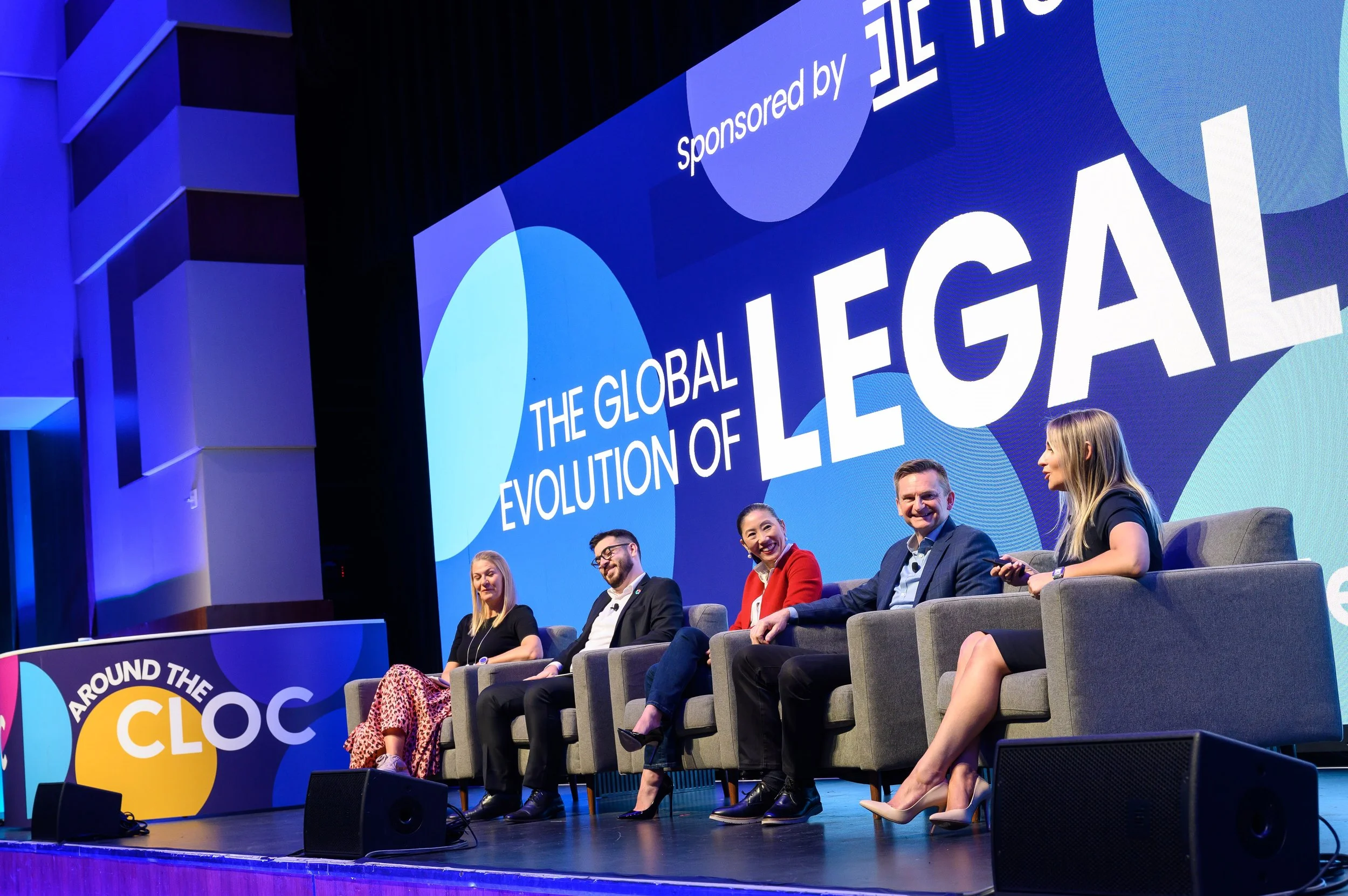 Panel discussion at a conference with five people seated on stage discussing the topic "The Global Evolution of Legal". The background features a display with the conference theme. The event appears to be sponsored, as mentioned in the backdrop.