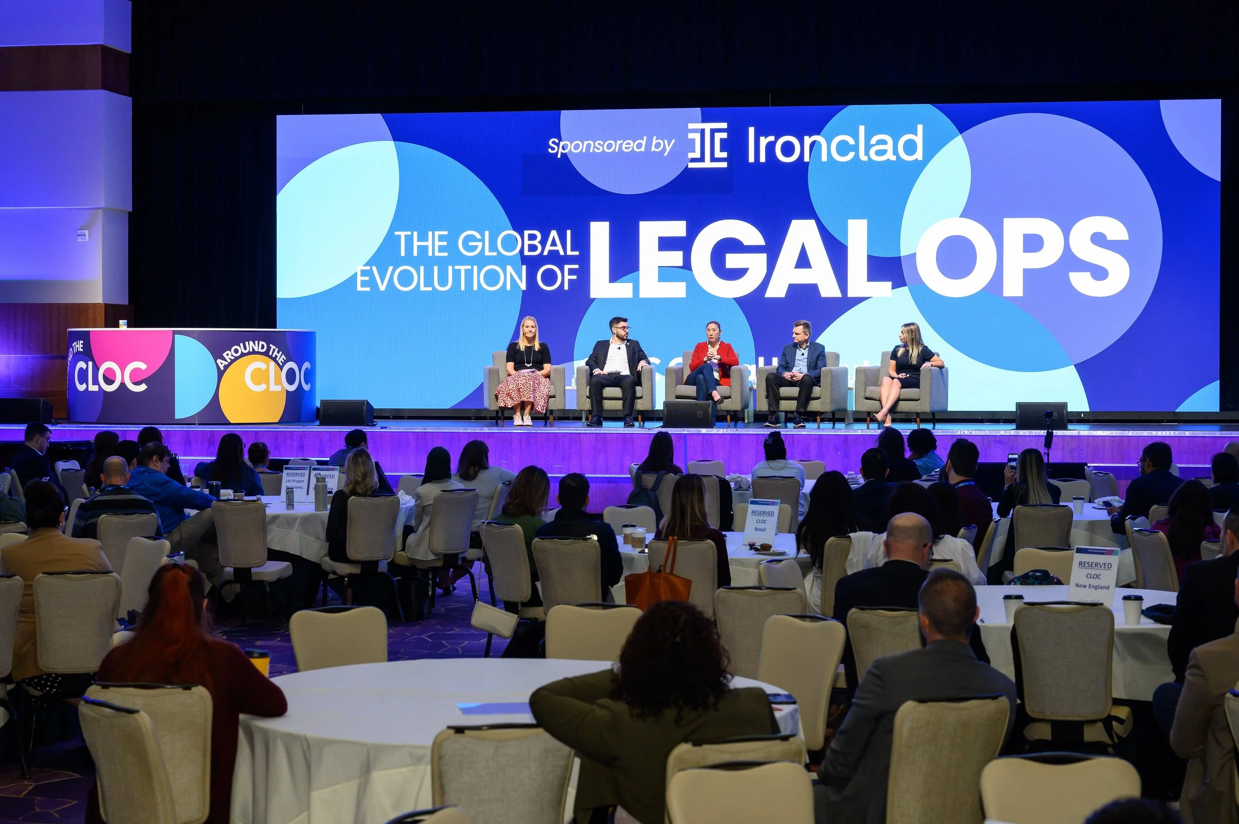 Panel discussion at a conference with "The Global Evolution of Legal Ops" displayed on a large screen, showing six panelists seated. The event is sponsored by Ironclad, with the audience seated at round tables.
