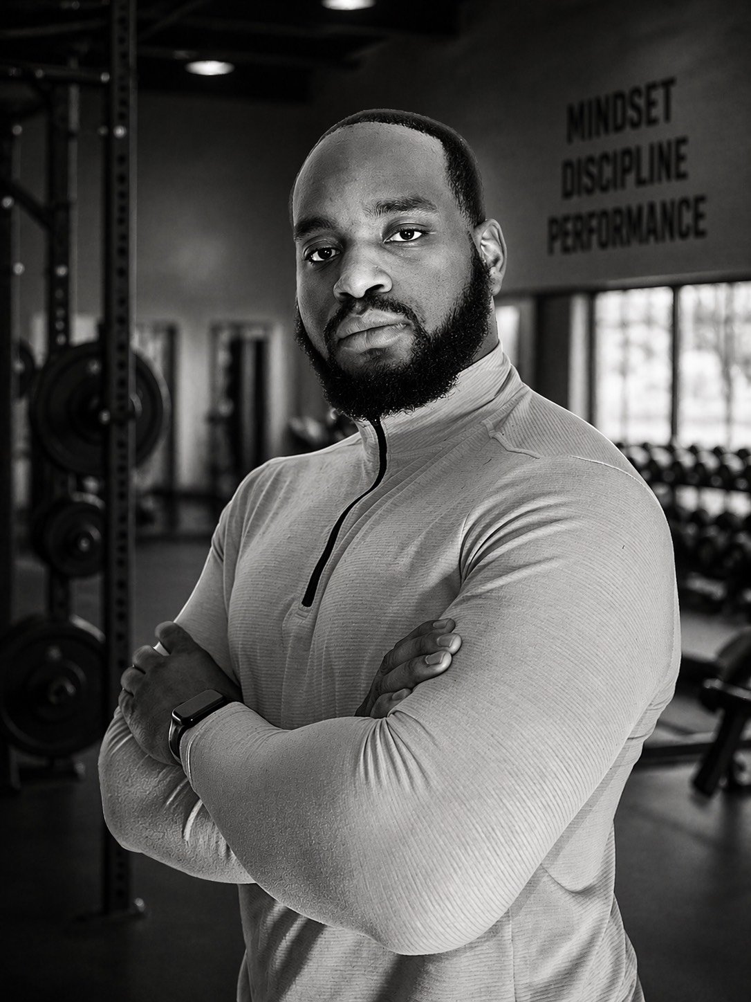 A man with a beard and short hair standing in a gym, looking at the camera with arms crossed. The background has gym equipment and a motivational quote on the wall reading "Mindset Discipline Performance."