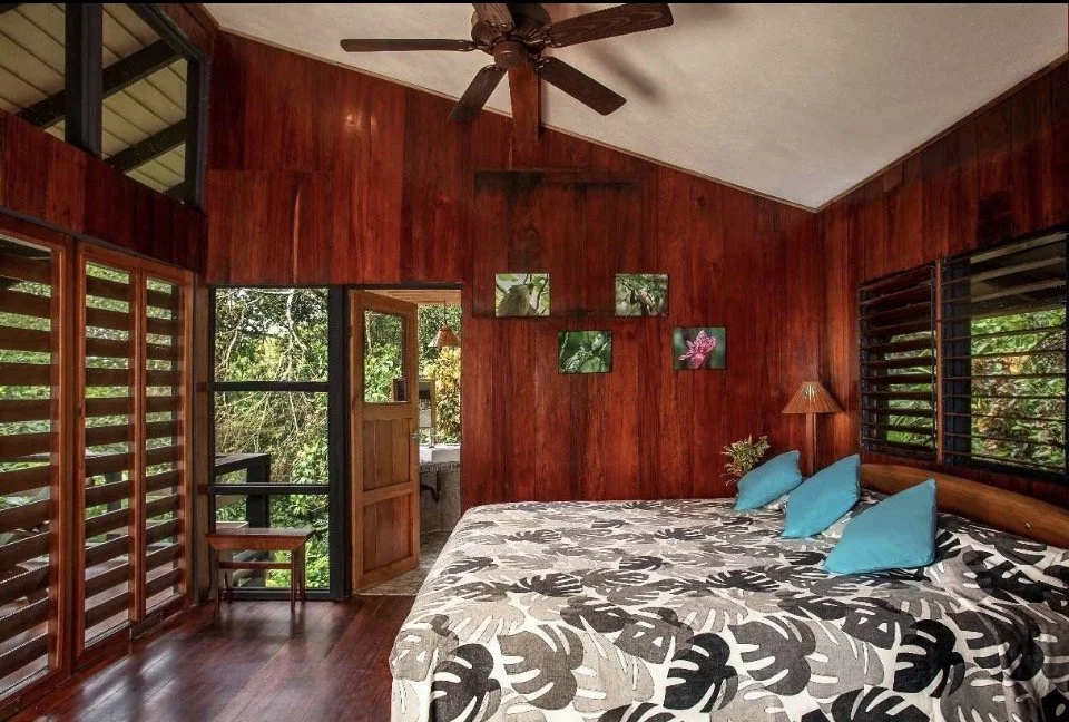 Room with wooden walls, large bed with floral bedding, four blue pillows, window slats, ceiling fan, small table and lamps, balcony with greenery outside.