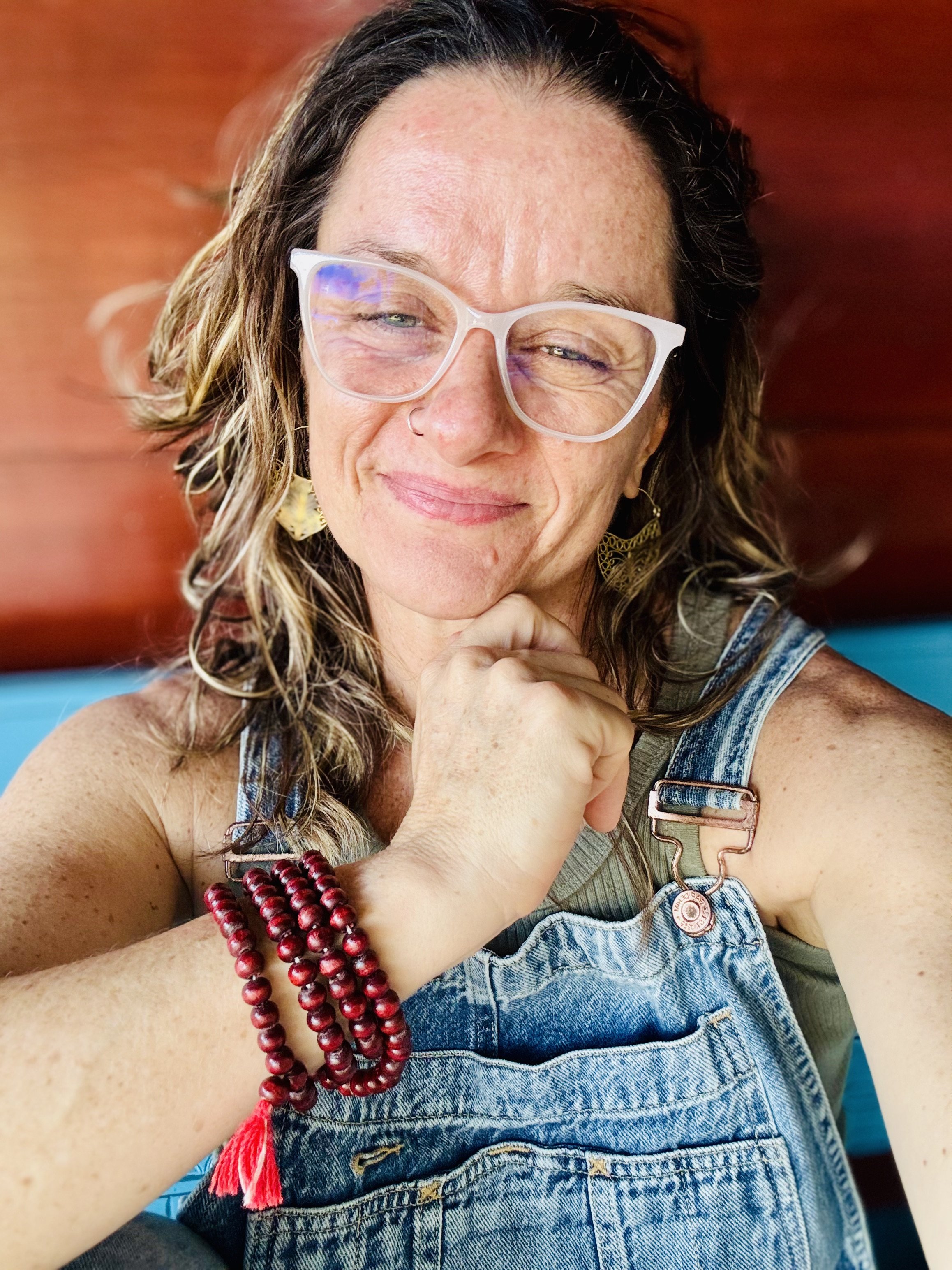 A woman with curly brown hair and freckles, wearing a green sleeveless top, denim overalls, clear glasses, and red bead bracelets, smiling with her eyes closed and resting her chin on her hand.