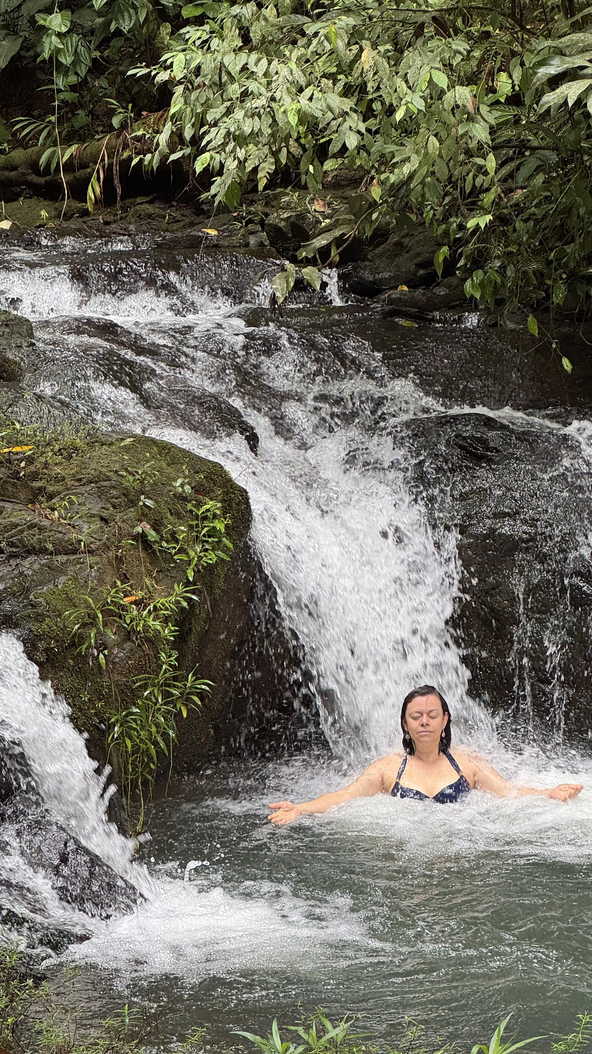 A woman with dark hair in a bathing suit relaxing in a small waterfall with lush green foliage in the background.