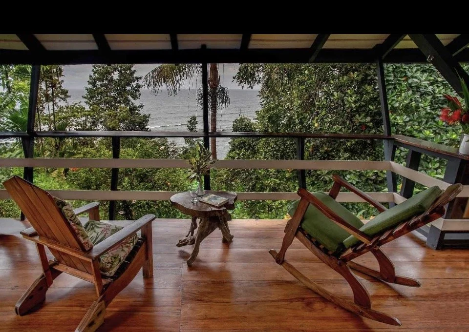 A cozy balcony with wooden furniture, including a rocking chair and a cushioned chair, overlooking lush greenery and the ocean.