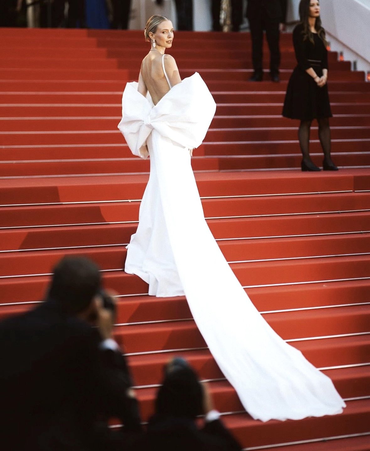 A woman in a white formal gown with a large bow on her back standing on red carpeted stairs at a formal event, with photographers and other attendees in the background.