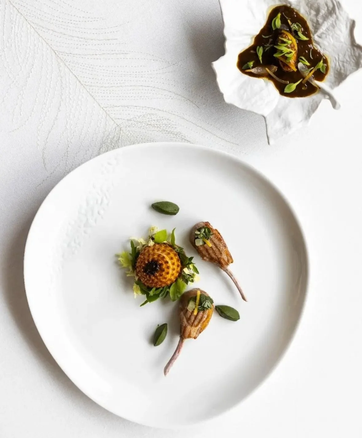 Gourmet dish with two small roasted vegetables, herbs, and a center element resembling a small honeycomb or textured sphere, presented on a white plate with green garnishes. A white textured cloth and a small dish with dark sauce and microgreens are visible in the background.