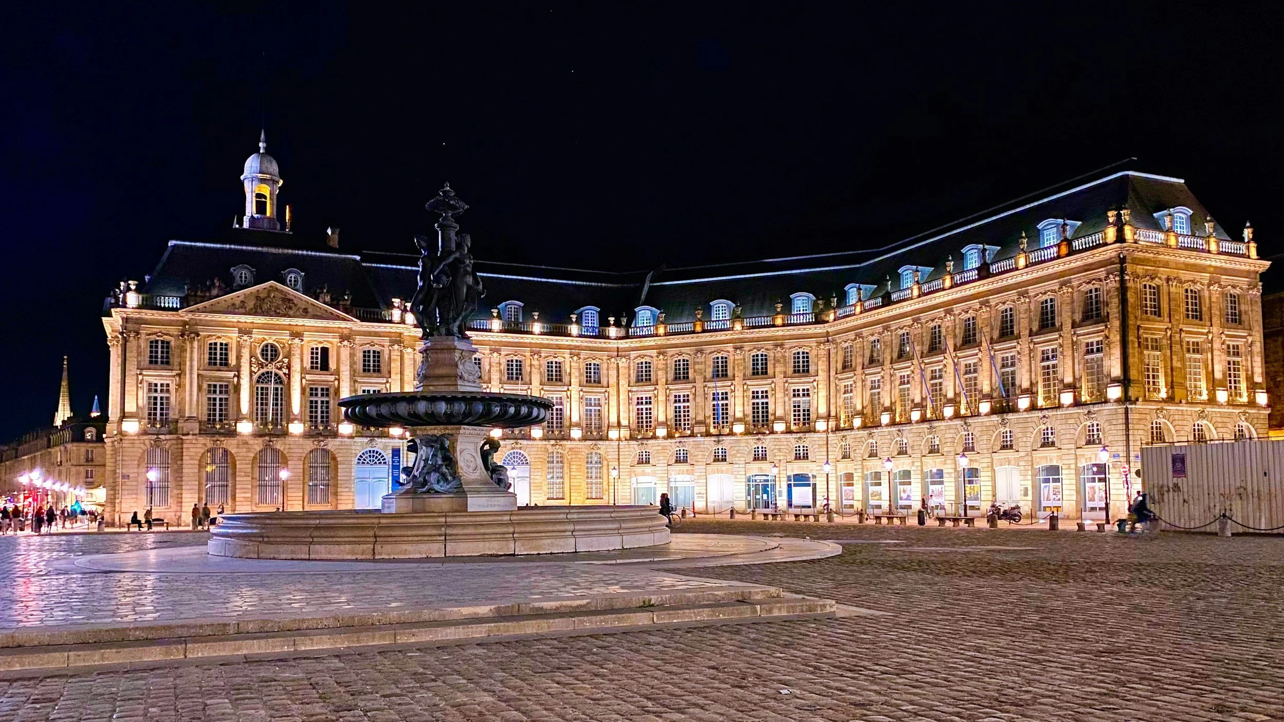 Bordeaux historic building illuminated at night, with a fountain in the foreground and cobblestone pavement, in a European city square.