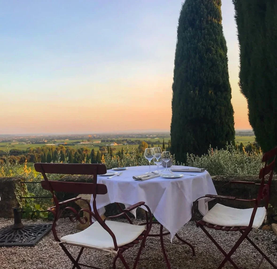 A table set for two with wine glasses, plates, and silverware on a white tablecloth, outdoors with tall cypress trees and a scenic view of the countryside during sunset.