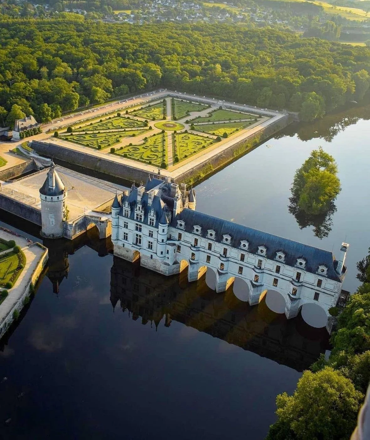 Aerial view of a historic castle surrounded by water with a formal garden and lush green forest in the background.