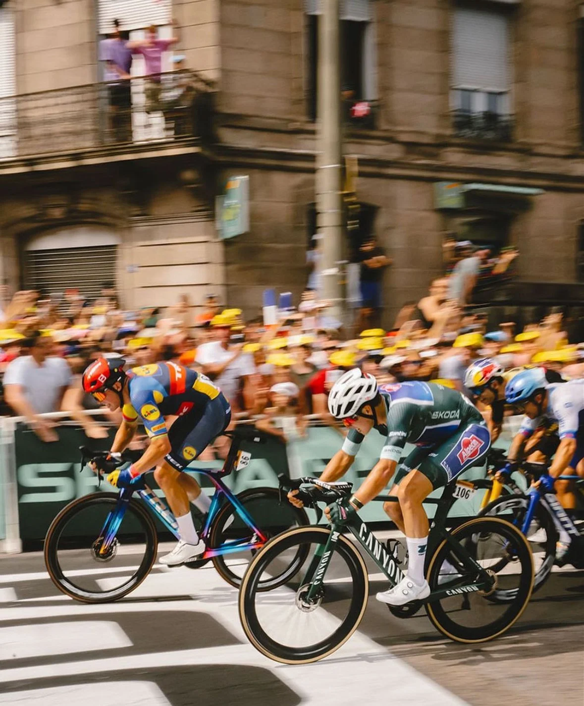 Cyclists racing in a crowded street with spectators watching from the sidewalk and building balconies.