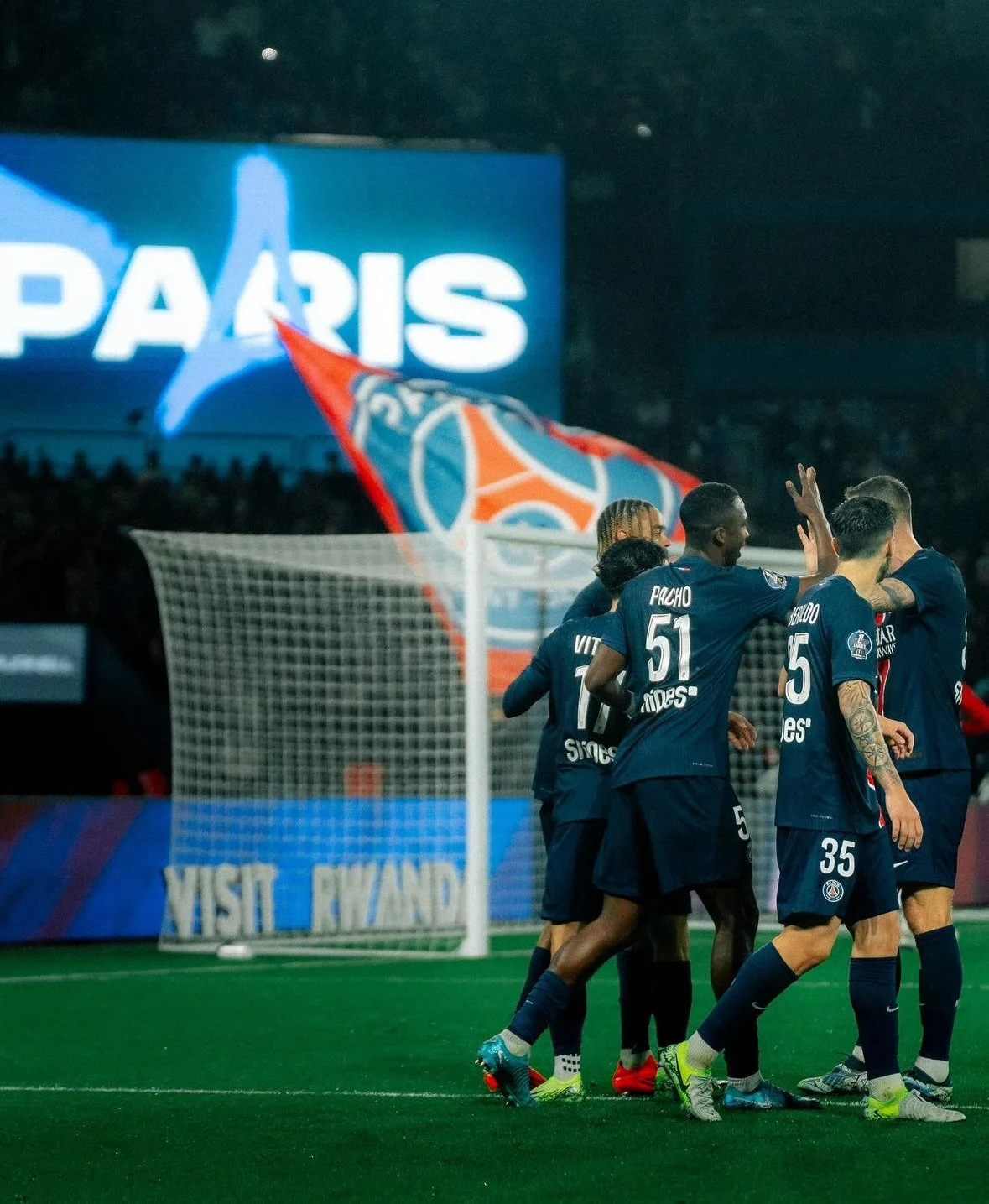 Soccer players in Paris Saint-Germain uniforms celebrating on the field, with a PSG flag and large illuminated PARIS sign in the background.