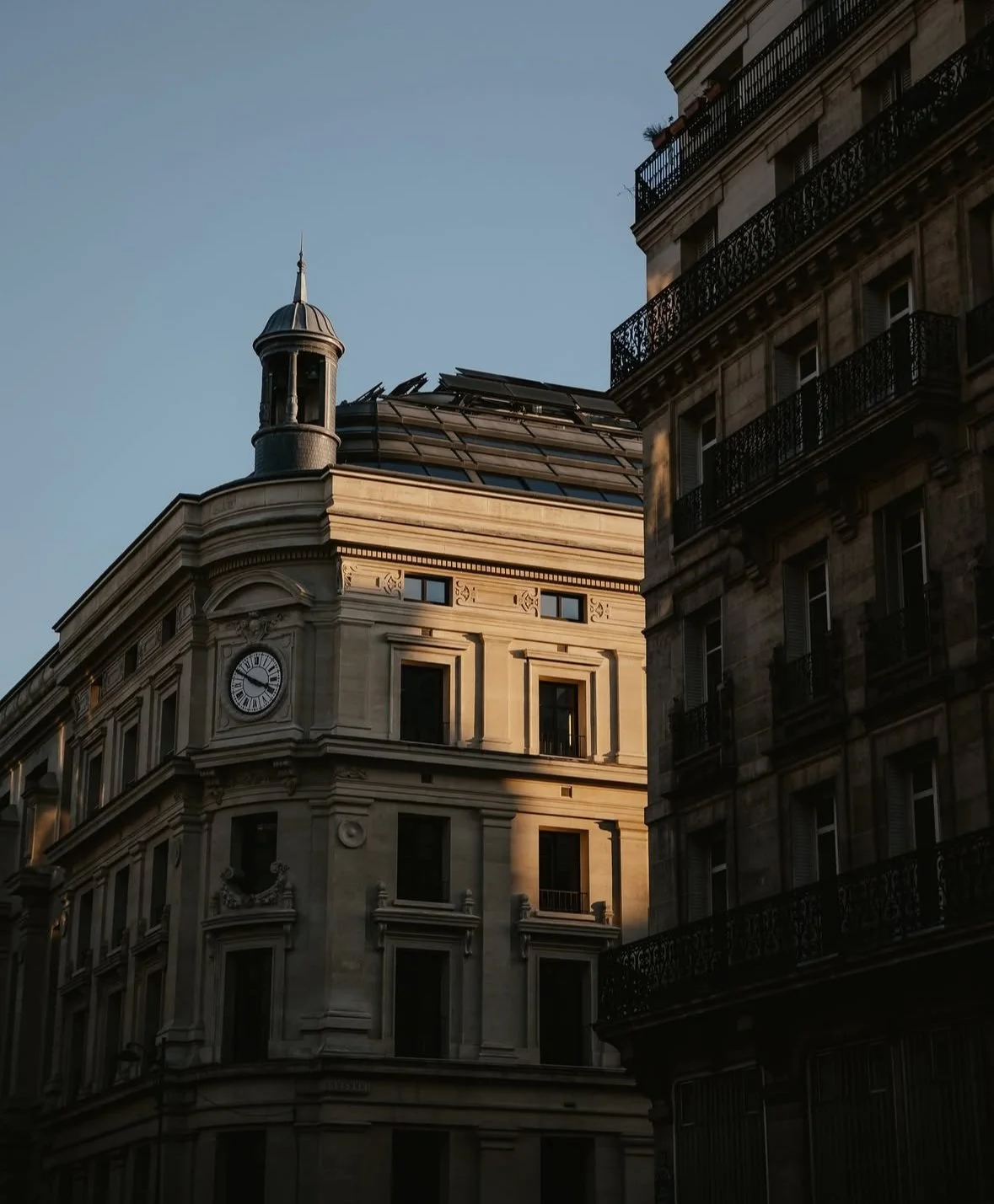 Sunlit historic building with a clock, ornate stone details, and a small tower at the top, next to a darker building with wrought iron balconies.