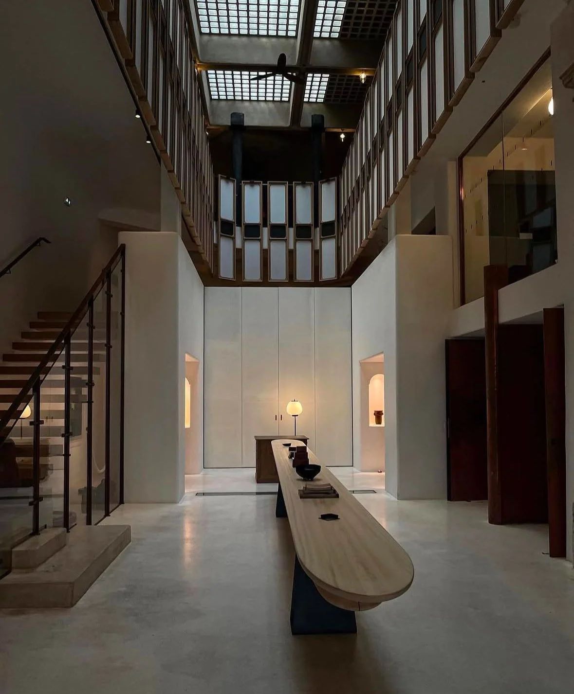 Interior of a modern, minimalistic lobby with a long wooden table, a lamp, and a staircase on the left. The upper level features a glass and wood balcony with grid-like ceiling skylights.