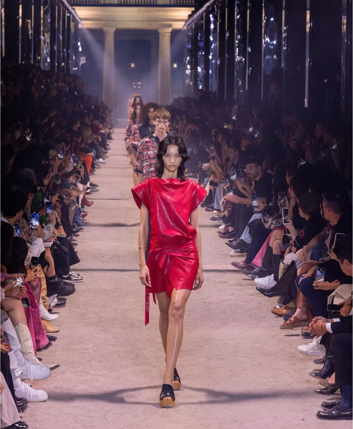 Models walking down a runway at a fashion show, with a female model in the foreground wearing a shiny red dress.