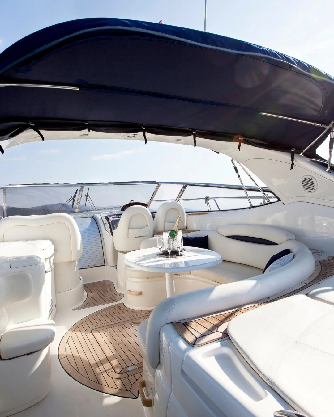 Interior of a yacht with a view of the water and distant mountains, featuring two white sofas, small white tables, and a deck with lounge chairs.