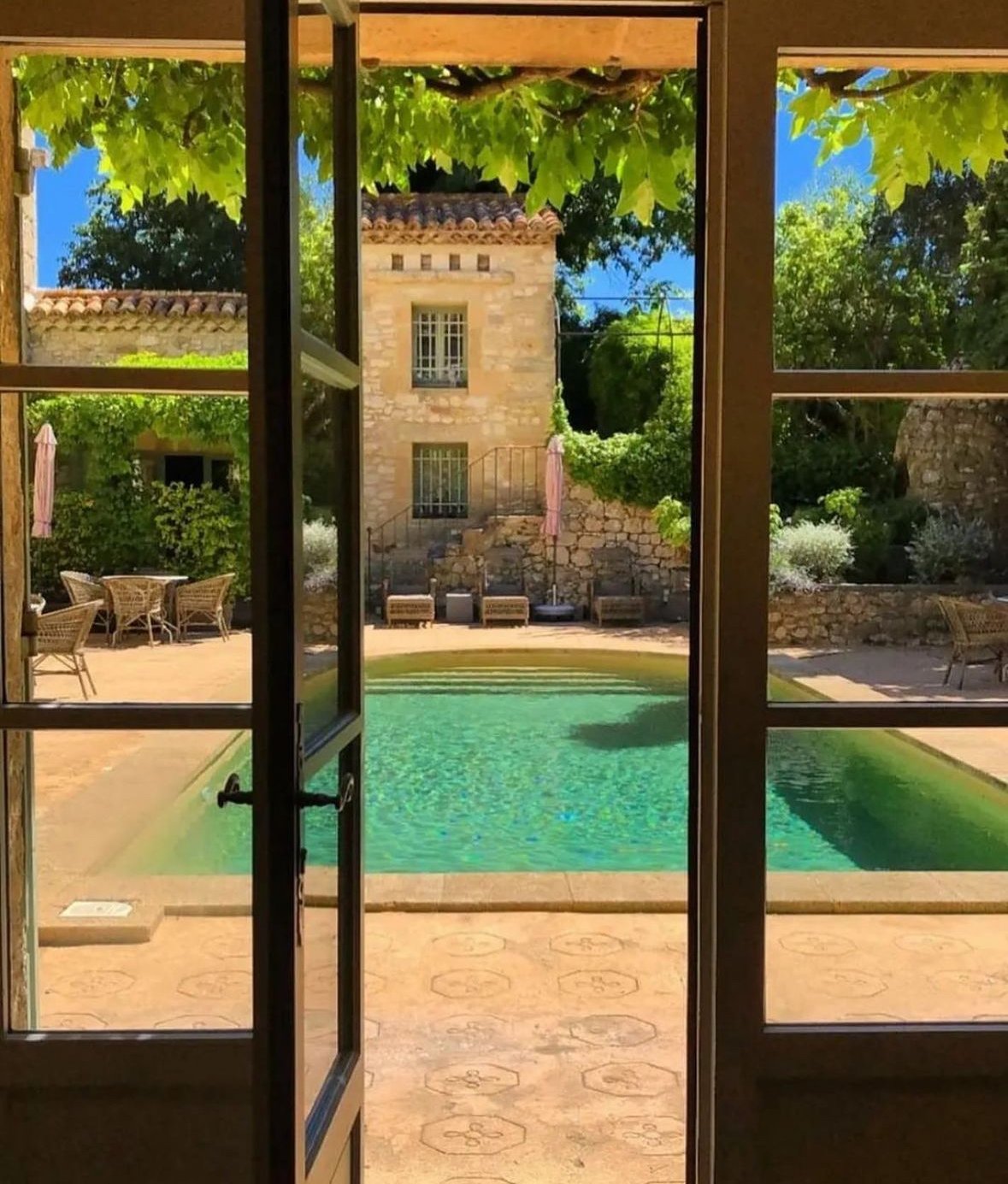 A view of a stone house with a tiled roof and windows, a swimming pool, and outdoor lounge chairs seen through an open door.