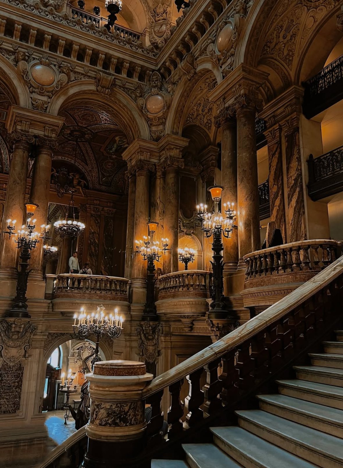 Elegant marble staircase inside a grand, ornate building with intricate gold and brown details, chandeliers, and marble columns.