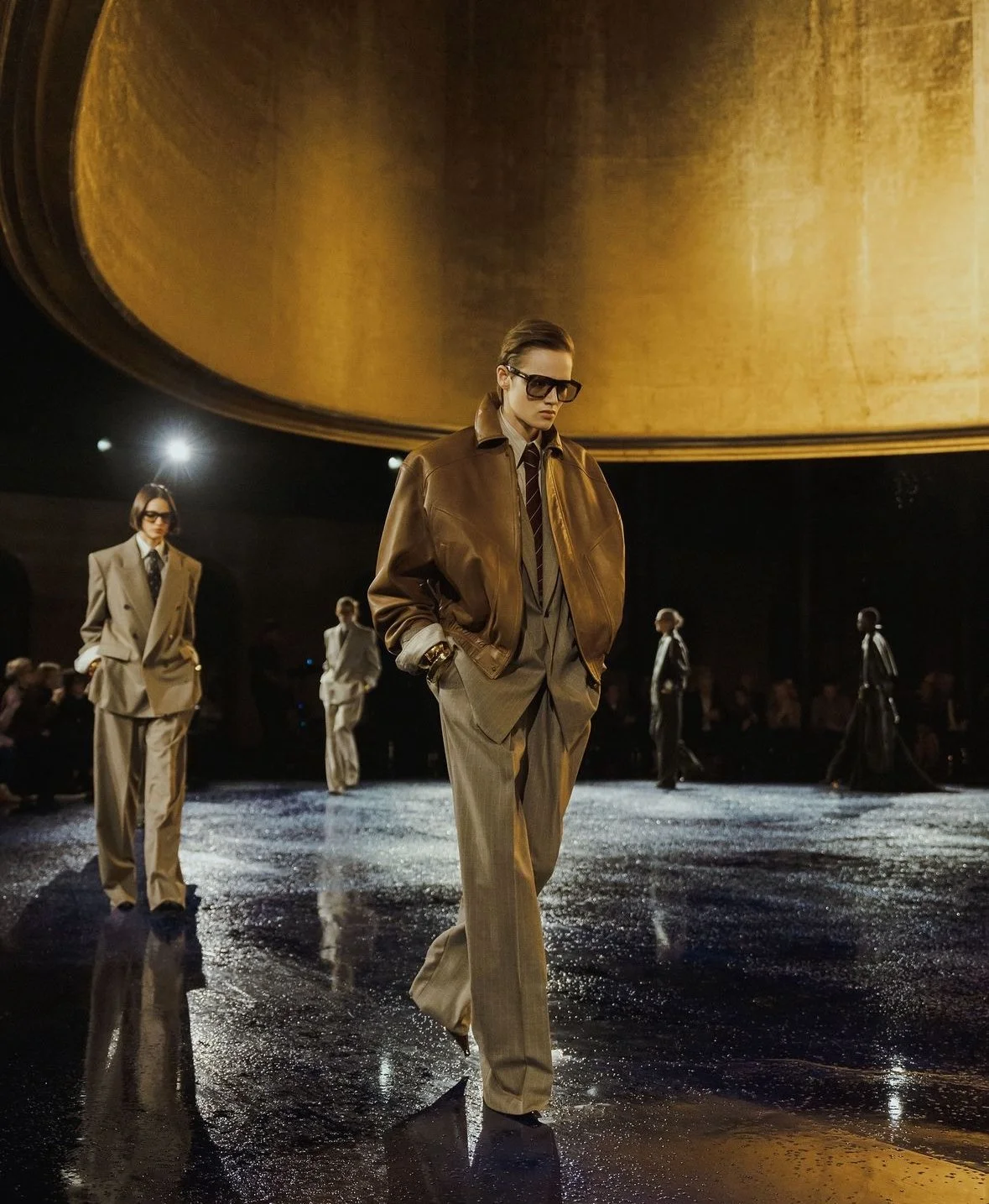 Models walking on a runway during a fashion show, wearing oversized suits and sunglasses, with a large golden circular backdrop.