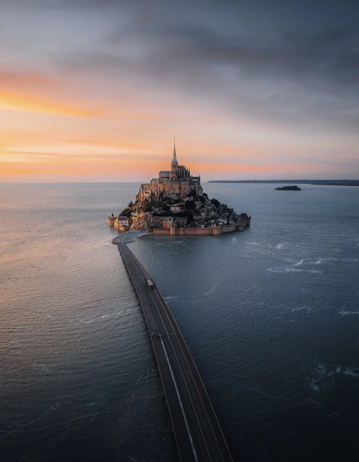 Aerial view of Mont Saint-Michel, an island commune in France with a historic abbey and castle, at sunset with a partly cloudy sky.