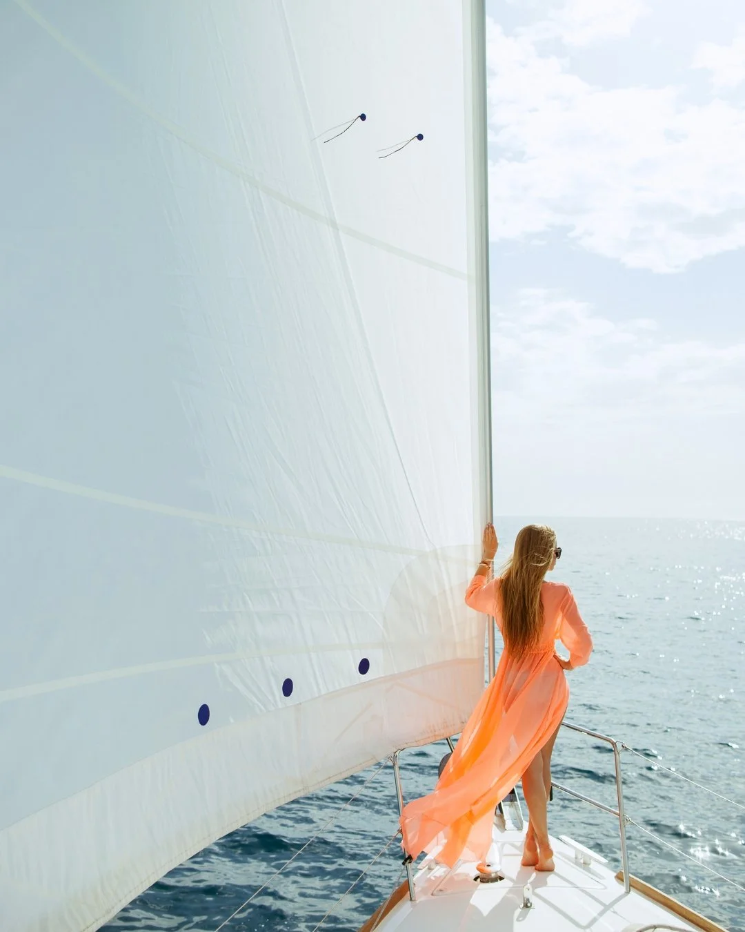 A woman in a flowing pink dress and sunglasses stands at the bow of a sailboat, holding onto the boat's mast, looking out at the open sea with a partly cloudy sky overhead.
