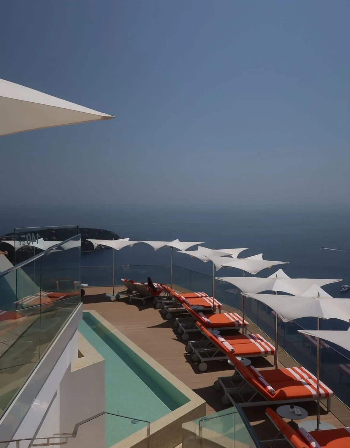 Poolside lounge chairs with orange cushions and white umbrellas on a balcony overlooking the ocean on a clear day.