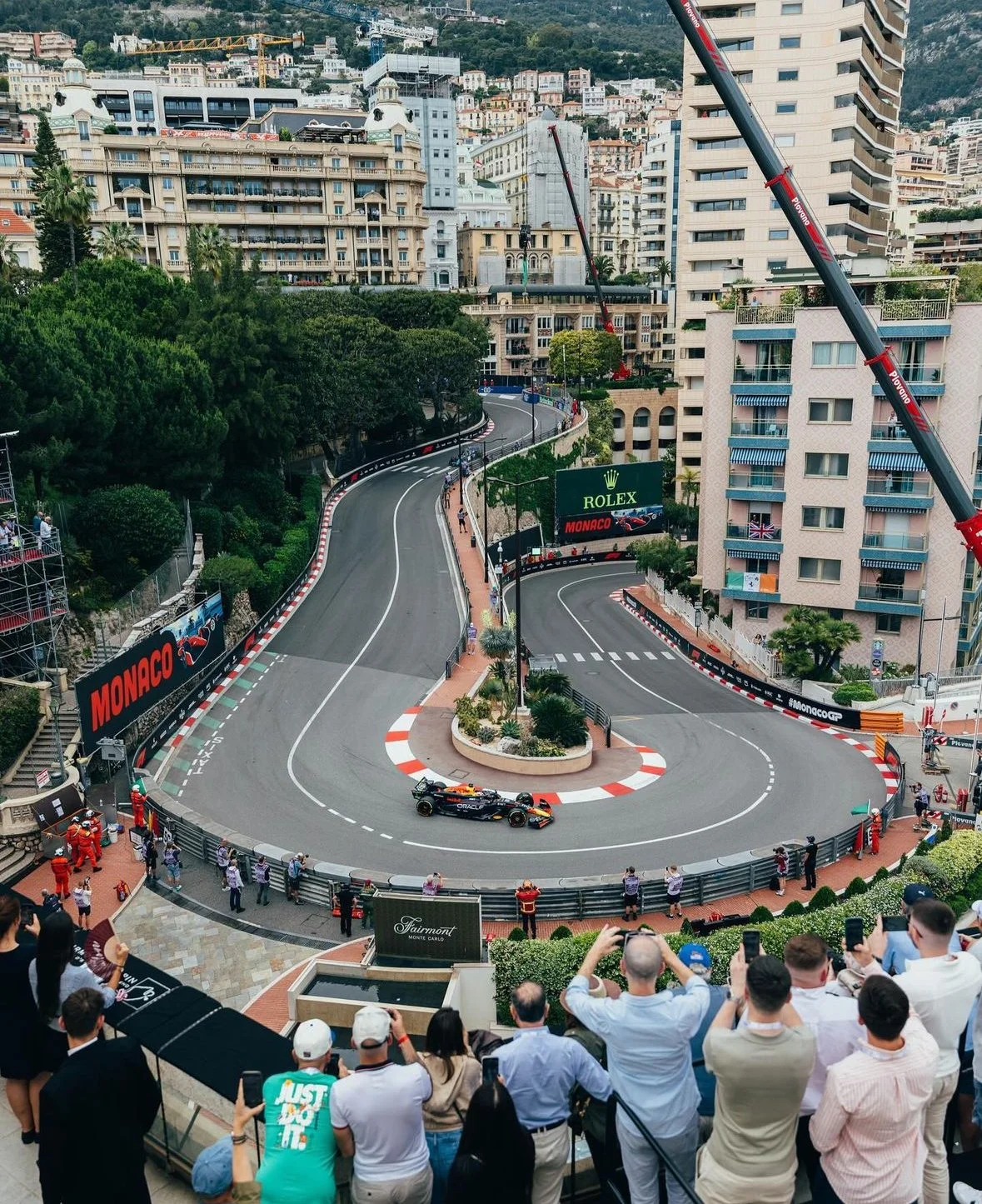 A Formula 1 car racing on a street circuit in Monaco, surrounded by spectators, tall buildings, and cityscape, with cranes and advertising banners for Rolex and Monaco Grand Prix.