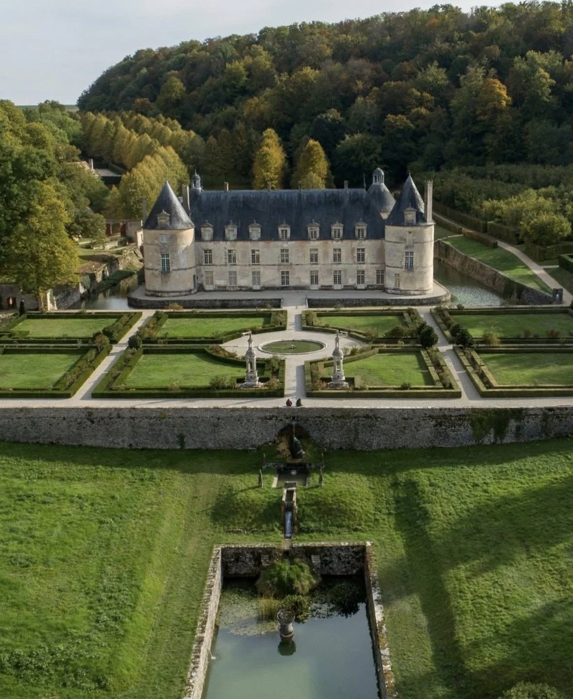 A historic castle with conical towers surrounded by a manicured garden and water features, set against green forested hills.