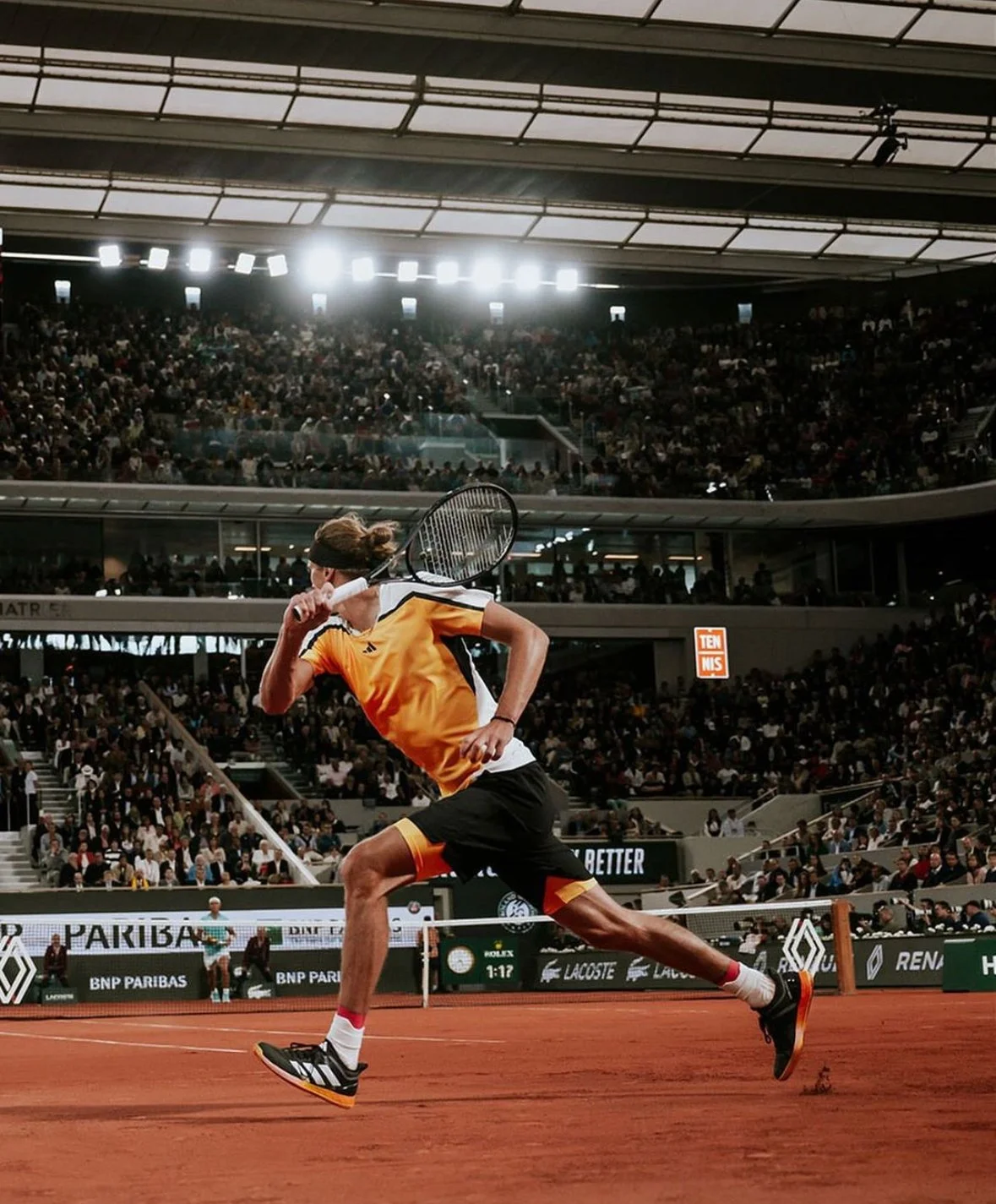 A tennis player in orange and black athletic clothing running on a clay court during a match, with a large audience in the stadium.