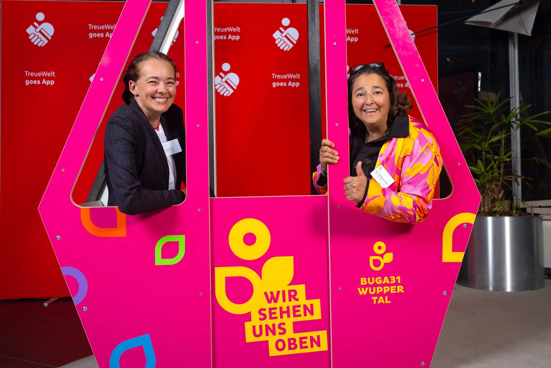 Zwei Frauen schauen aus einer pinkfarbenen Gondel, eine lächelnd und die andere zeigt Daumen hoch. Der Hintergrund hat einen roten Bereich mit weißem Logo und Text. Die Gondel hat bunte Designs und Schriftzüge, darunter 'WIR SEHEN UNS OBEN' und Ortsa