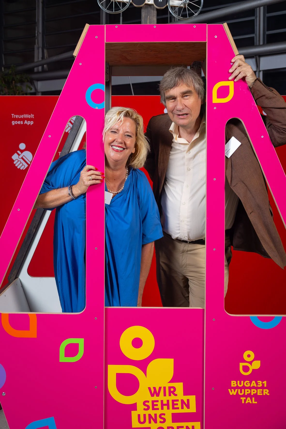 Ein Mann und eine Frau stehen in einer Gondel in einer Erlebniswelt, die rosa gefärbt ist, und lachen. Im Hintergrund sind bunte Logos und ein roter Hintergrund mit Text in deutscher Sprache.