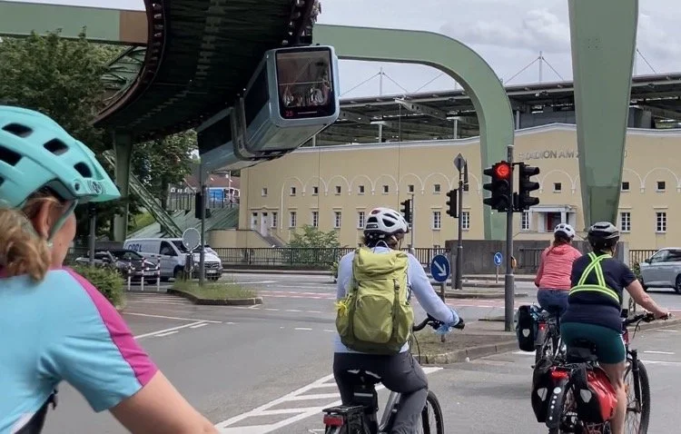 Das Bild zeigt eine Gruppe von Radfahrenden. Sie stehen an einer roten Ampel am Stadion am Zoo in Wuppertal. Über der Gruppe fährt die Wuppertaler Schwebebahn.