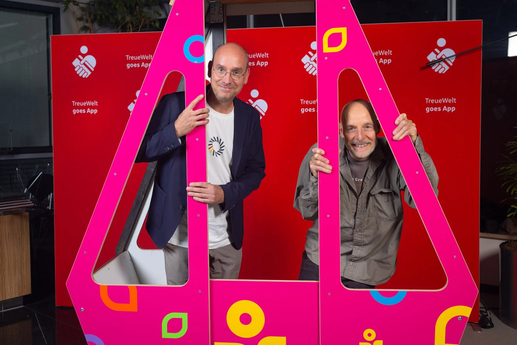 Zwei Männer schauen durch große pinke Buchstaben an einer roten Wand, mit einem Hintergrund der Anzeige 'TreueWelt goes App'.