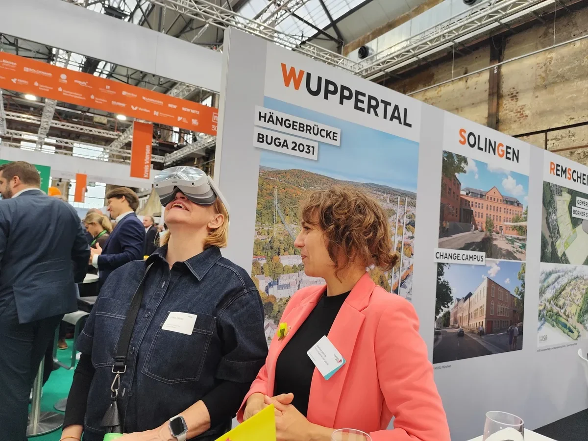 Dieses Bild zeigt eine Frau, die auf der polis convention in Düsseldorf eine VR-Brille trägt und begeistert ist über die Möglichkeit, schon heute auf der ersten urbanen Hängebrücke der Welt stehen zu können, die für BUGA31 Wuppertal geplant ist.