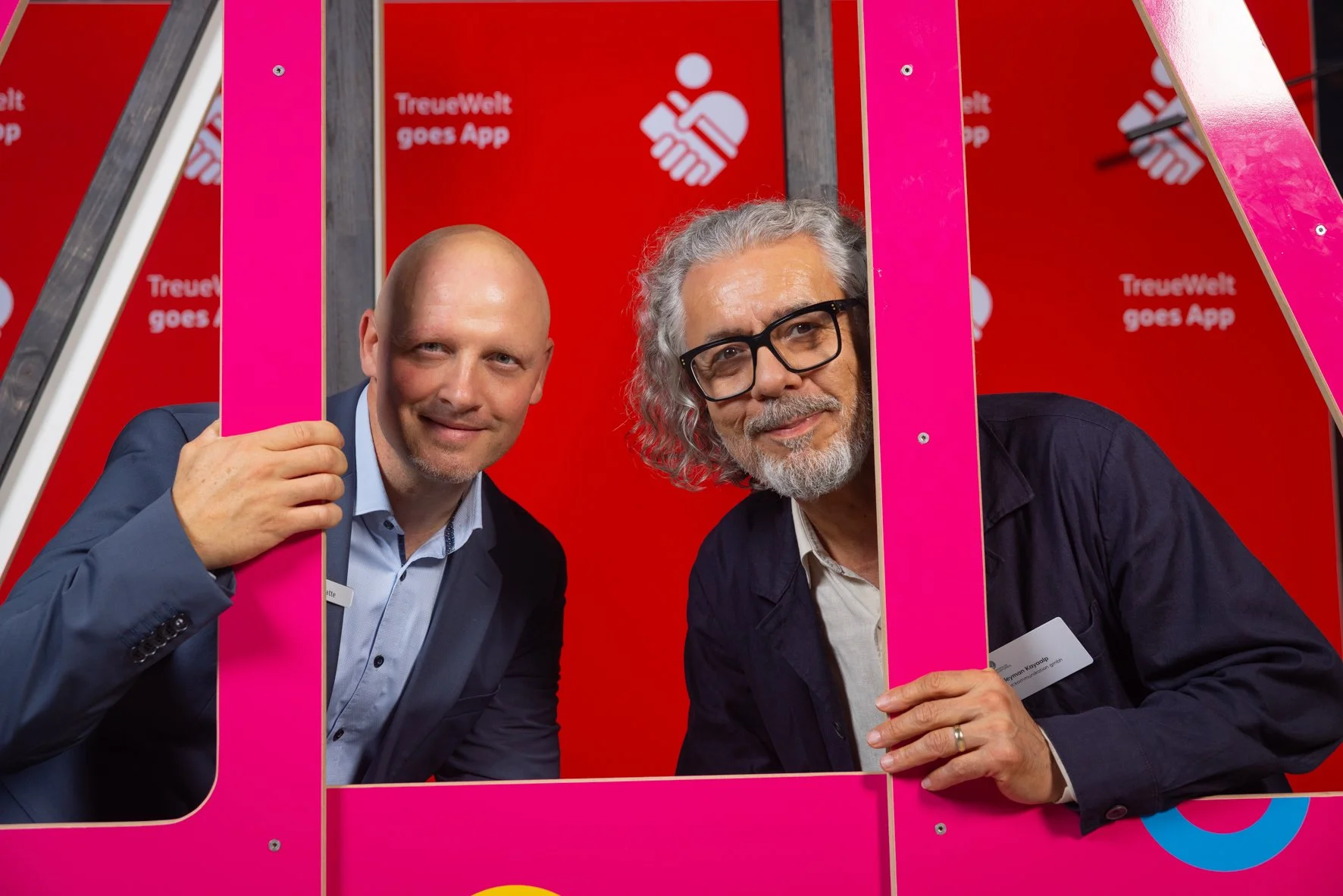 Zwei Männer posieren hinter einer pinken Rahmenkonstruktion vor einem roten Hintergrund mit weißem Logo und Text, das eine Hand und die Worte 'Treuwelt goes App' zeigt. Einer hält die Rahmenkonstruktion, beide schauen in die Kamera und lächeln.