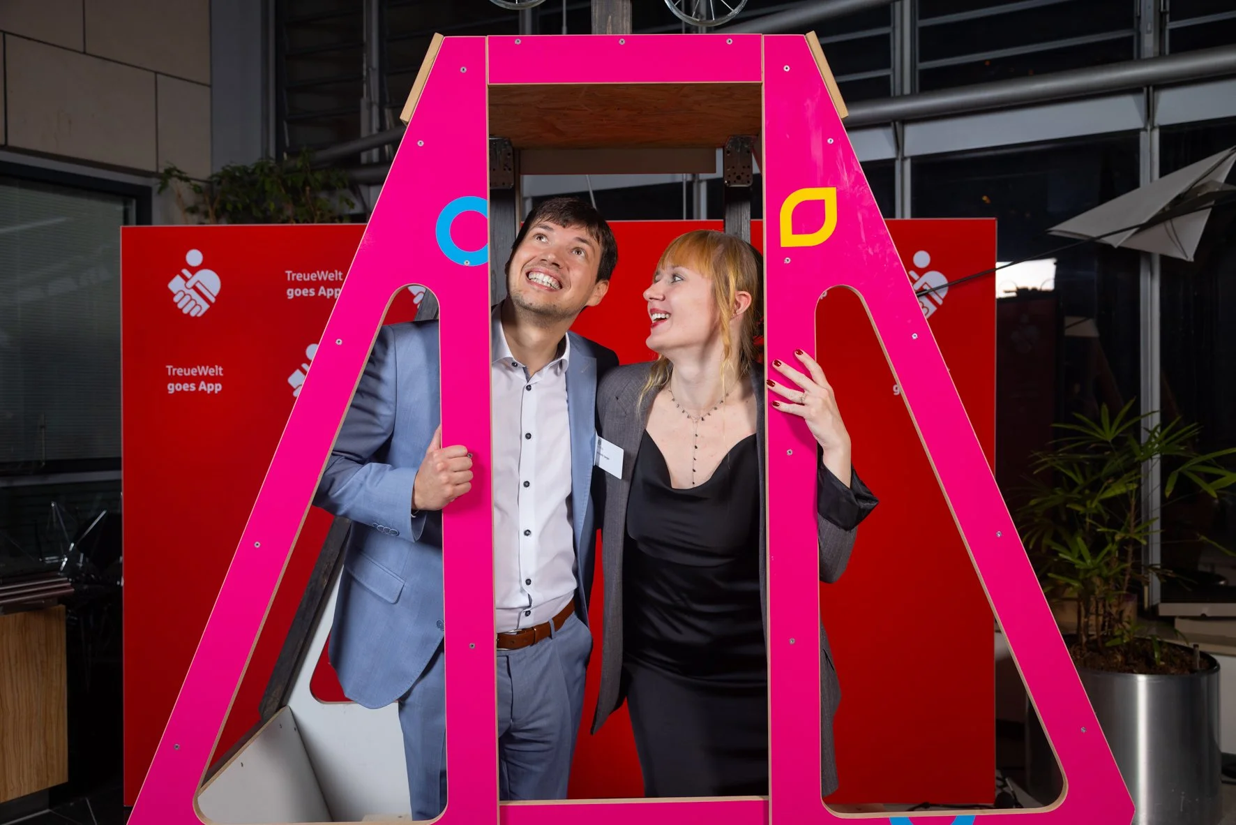 Zwei lachende Menschen, ein Mann im grauen Anzug und eine Frau in dunkler Kleidung, stehen in einem pinken Spielplatzgerüst bei einer Veranstaltung.