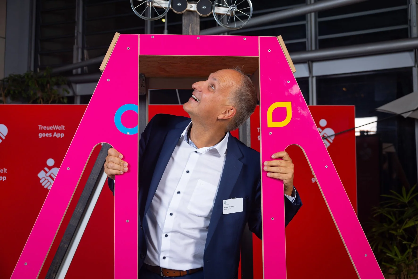 Ein Mann in einem dunklen Anzug steht hinter einer pinkfarbenen, großen Buchstabenform. Er schaut nach oben und lächelt. Im Hintergrund sind rote Werbebanner und ein moderner Gebäudeteil.