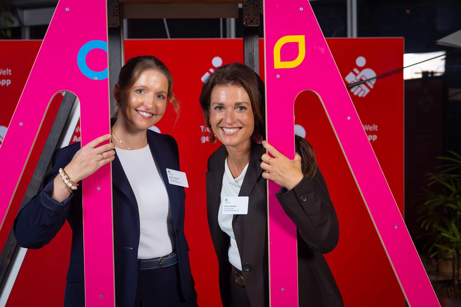 Zwei Frauen posieren hinter einer pinkfarbenen Rahmenstruktur bei einer Veranstaltung. Beide tragen dunkle Blazer und tragen Namensschilder.