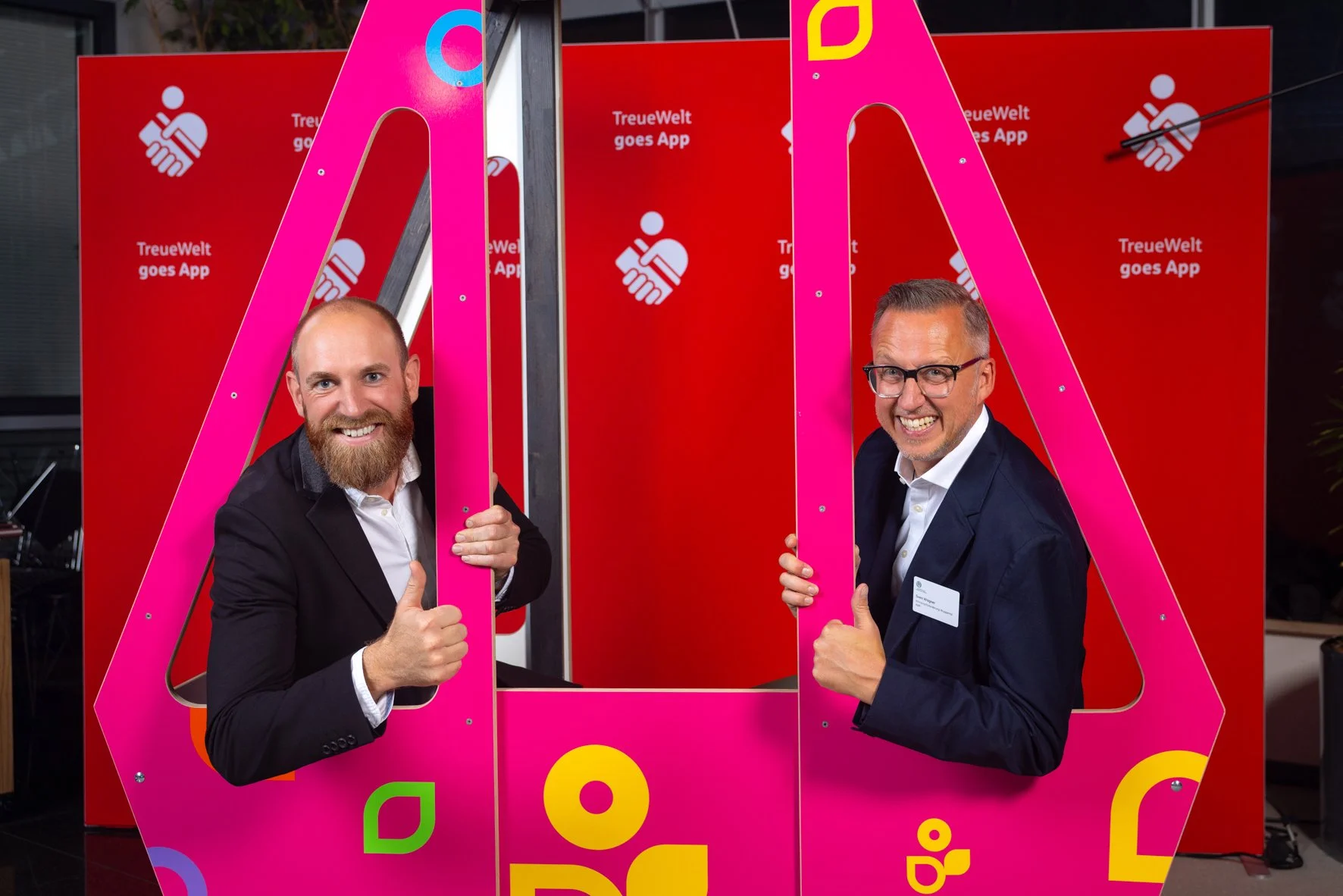 Zwei Männer in Anzügen posieren hinter pinken Stangen, die eine große, rot Hintergrundtafel mit dem Text 'TreueWelt goes App' und einem Handshake-Logo im Hintergrund halten.