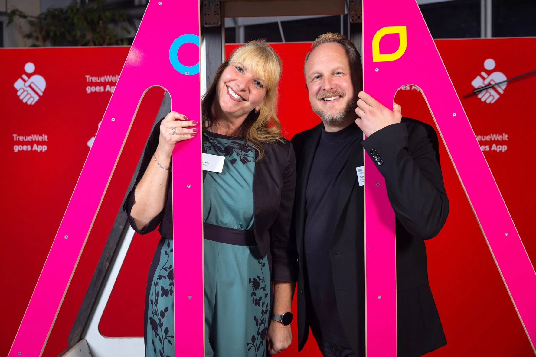 Zwei lächelnde Menschen, eine Frau und ein Mann, posieren hinter pinkfarbenen, großen Buchstaben vor einem roten Hintergrund, der das Logo und den Text "TreueWelt goes App" zeigt.