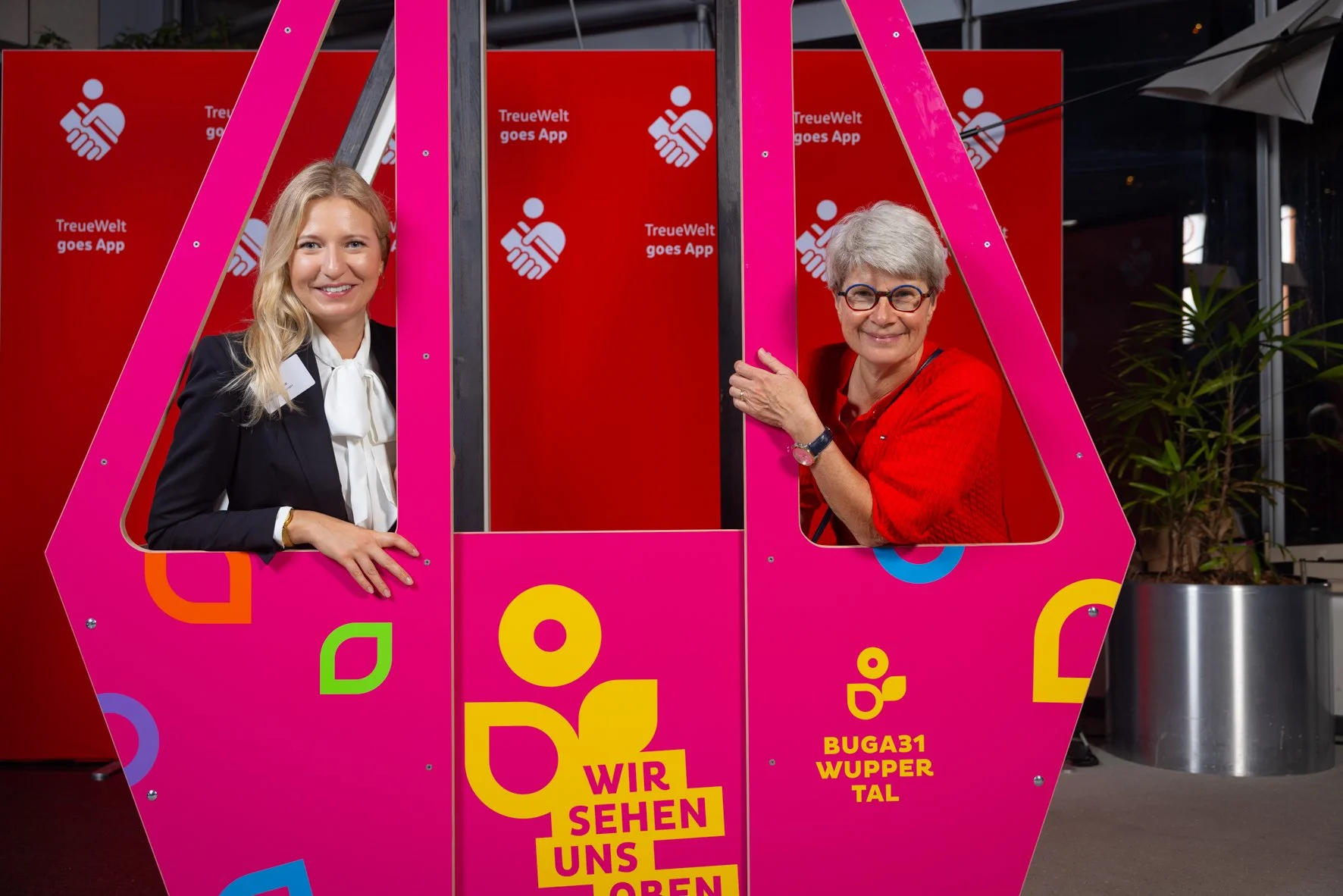 Zwei Frauen, eine jüngere und eine ältere, posieren in einem pinkfarbenen Fotokabinenrahmen mit bunten Designs. Im Hintergrund befindet sich ein roter Bildschirm mit weißen Texten, die von einer App namens TreueWelt werben. Die jüngere Frau hat blond