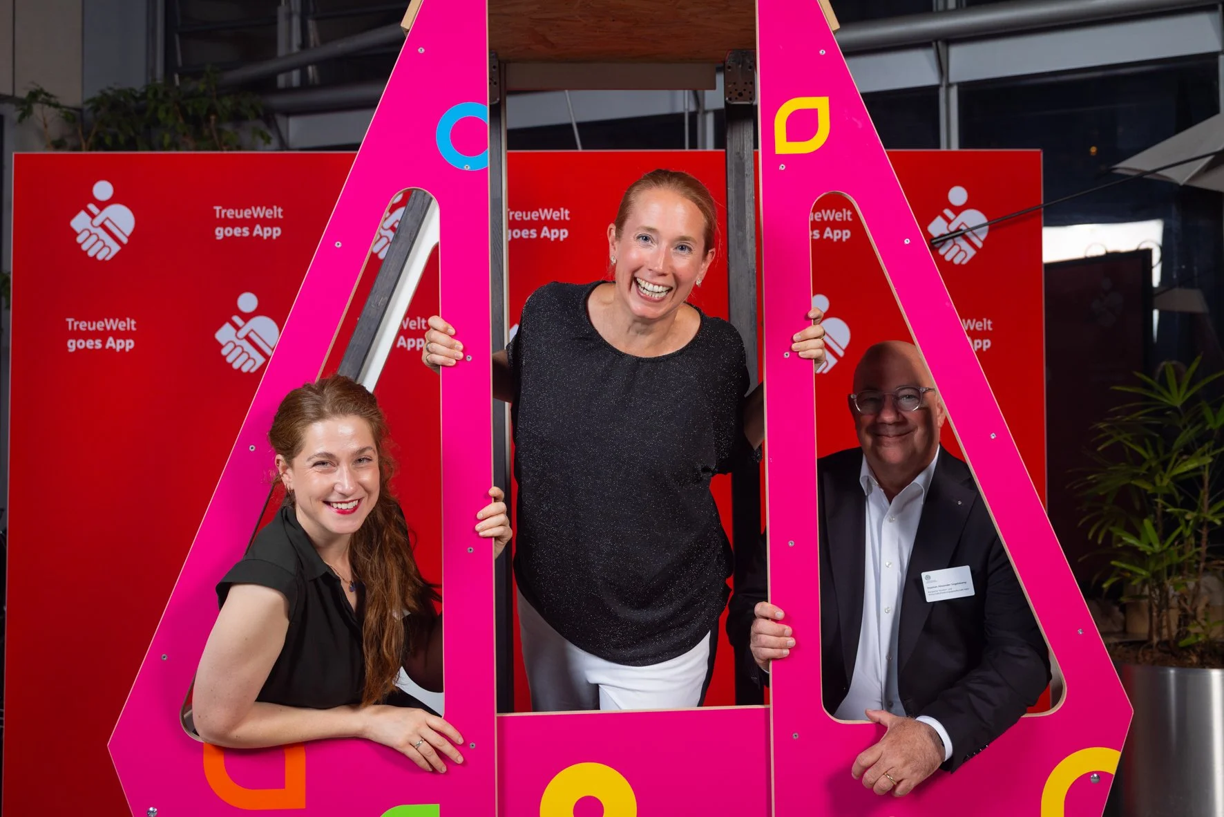 Drei Personen posieren hinter einer großen pinken Rahmen bei einer Veranstaltung, vor einem roten Hintergrund mit weißen Text und Logo.