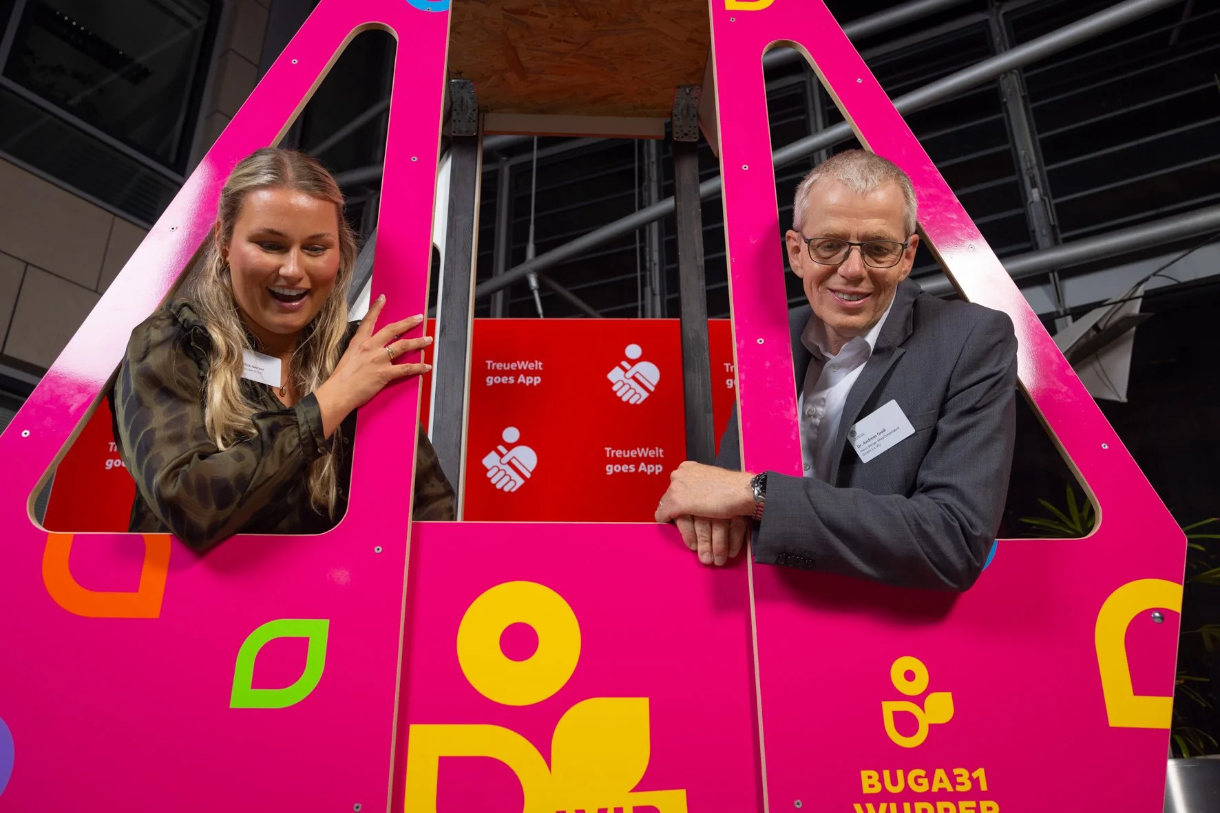 Ein Mann und eine Frau, die aus einem pinkfarbenen, plakatbedeckten Fahrzeug herausschauen, während sie bei einer Veranstaltung zusammen lachen. Im Hintergrund sind rote Tafeln mit weißen Symbolen und der deutschen Beschriftung 'TreueWelt goes App' s