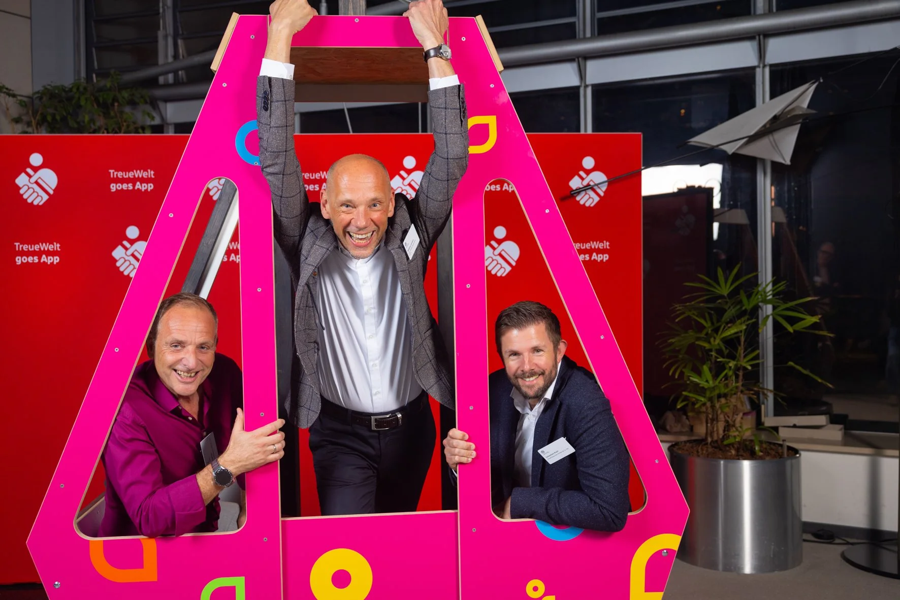 Drei Männer posieren mit einem pinken Rahmen und einem kleinen Dach oben, im Hintergrund rote Banner mit Logo und Text. Einer der Männer hebt die Arme in die Luft, alle lächeln.