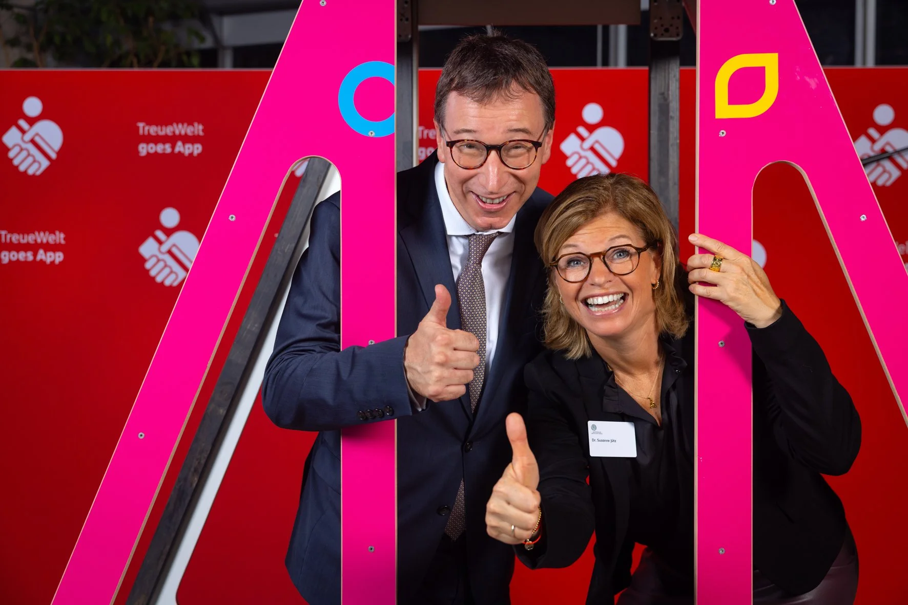 Zwei lachende Menschen, ein Mann und eine Frau, zeigen Daumen hoch, stehen hinter einer großen pinkfarbenen Buchstabe 'A' bei einer Veranstaltung mit einem roten Hintergrund, der das Logo der TreueWelt App zeigt.