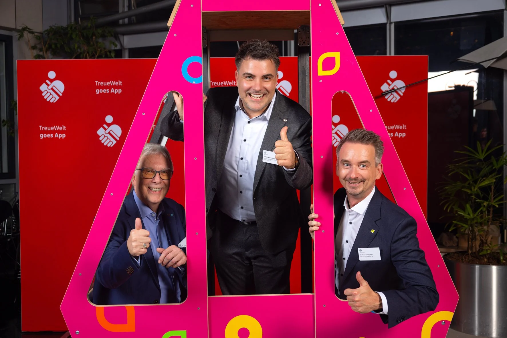 Drei Männer in formeller Kleidung posieren hinter einem pinkfarbenen Rahmen in Form eines Buchstaben A. Sie zeigen Daumen hoch und lächeln bei einer Veranstaltung mit einem roten Hintergrund, der die Aufschrift 'TreueWelt goes App' und Logos enthält.