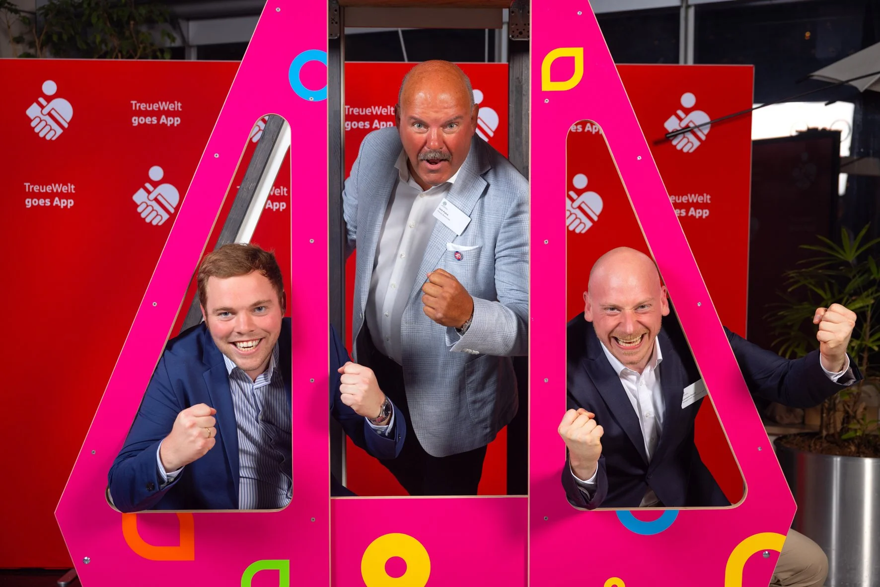 Drei Männer posieren fröhlich hinter pinken Rahmen, mit roten Hintergründen mit weißen Symbolen und Text, der 'TreueWelt goes App' sagt.