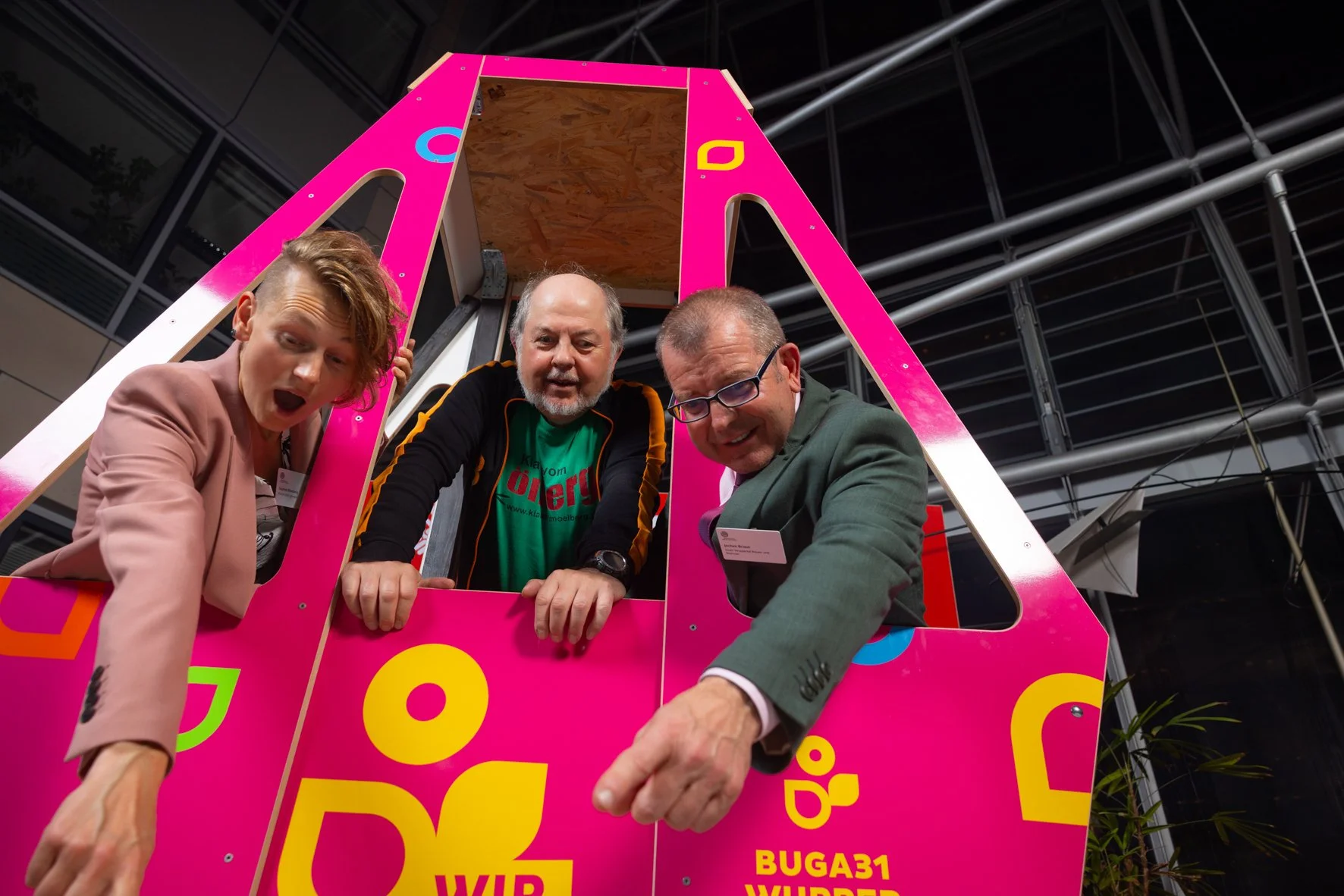 Drei Menschen stehen in einer pinkfarbenen, eckigen, kreativ gestalteten Gondel, die mit bunten Buchstaben und Zahlen dekoriert ist. Sie schauen nach unten, während sie die Gondel berühren oder darin stehen, in einem modernen Gebäude mit sichtbarer D