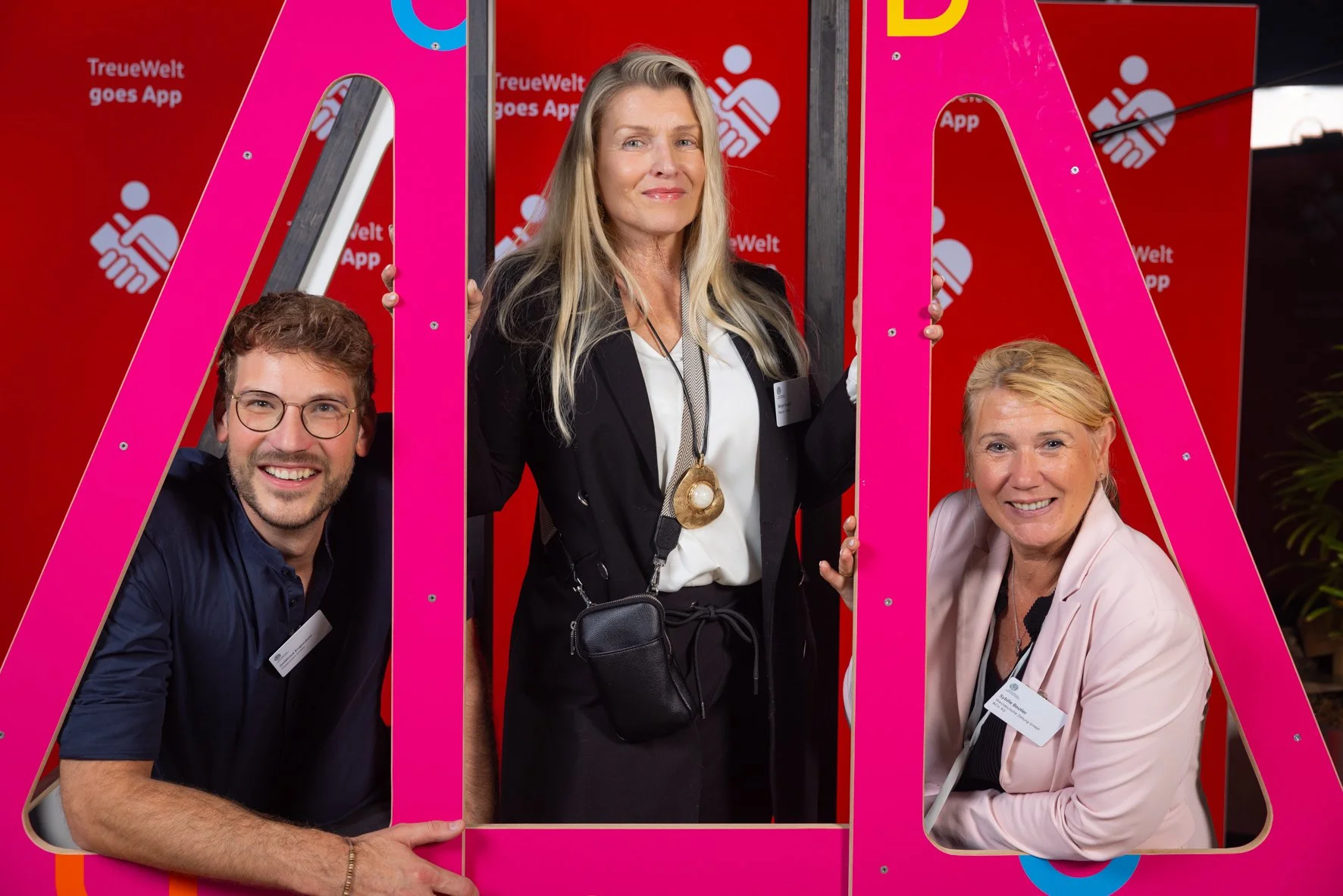 Drei Menschen posieren in einer bunte Fotokabine. Der Hintergrund ist rot mit weißen Symbolen und Text. Die Frau in der Mitte trägt eine schwarze Jacke mit großen Ketten und eine schwarze Tasche, die anderen beiden, ein Mann und eine Frau, tragen hel